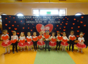 Grupa dzieci unosi przed sobą serca, w tle dekoracja na Dzień Babci i Dziadka.